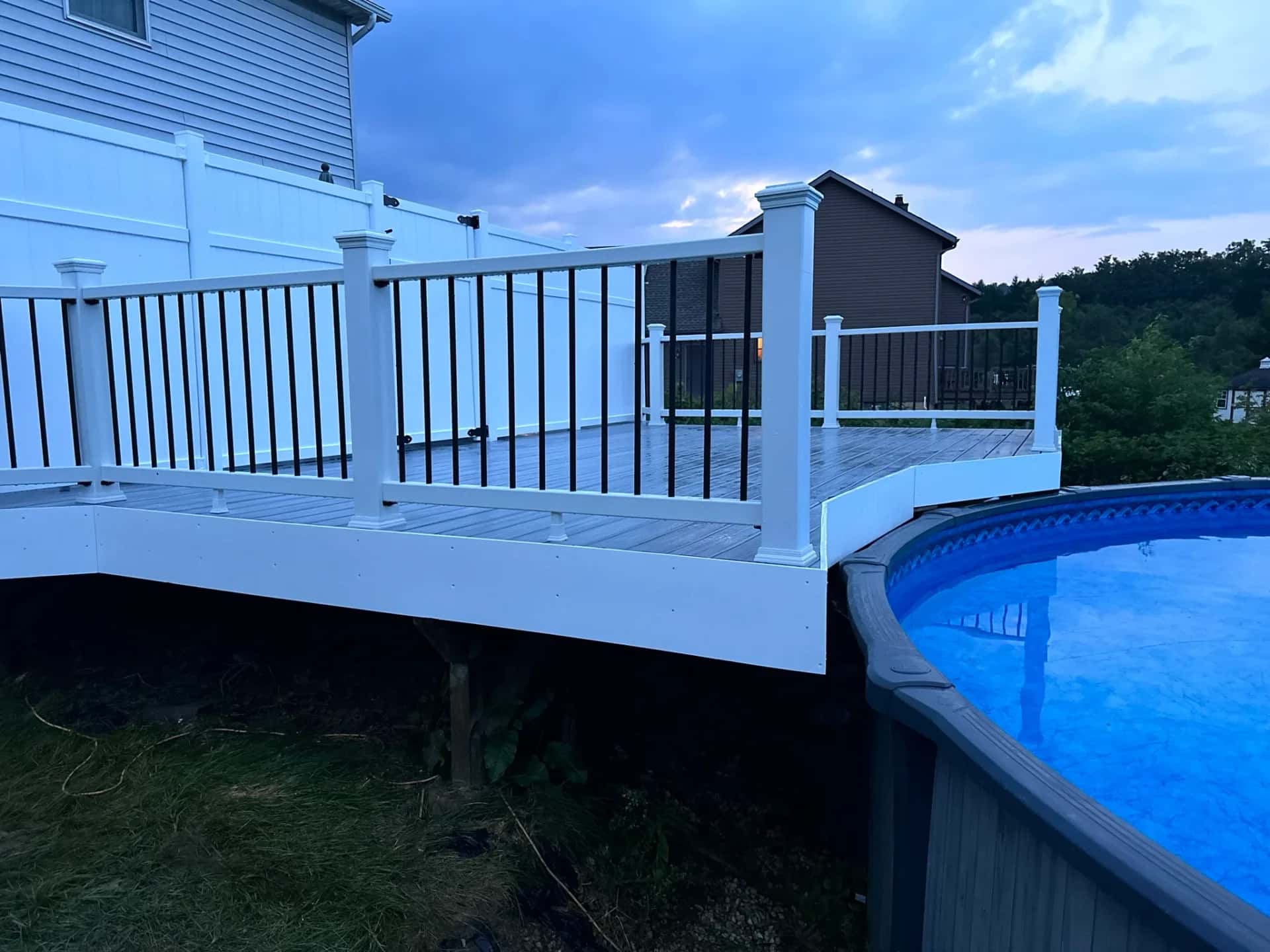 Gray composite pool deck with white railing and black balusters at dusk beside an above-ground pool
